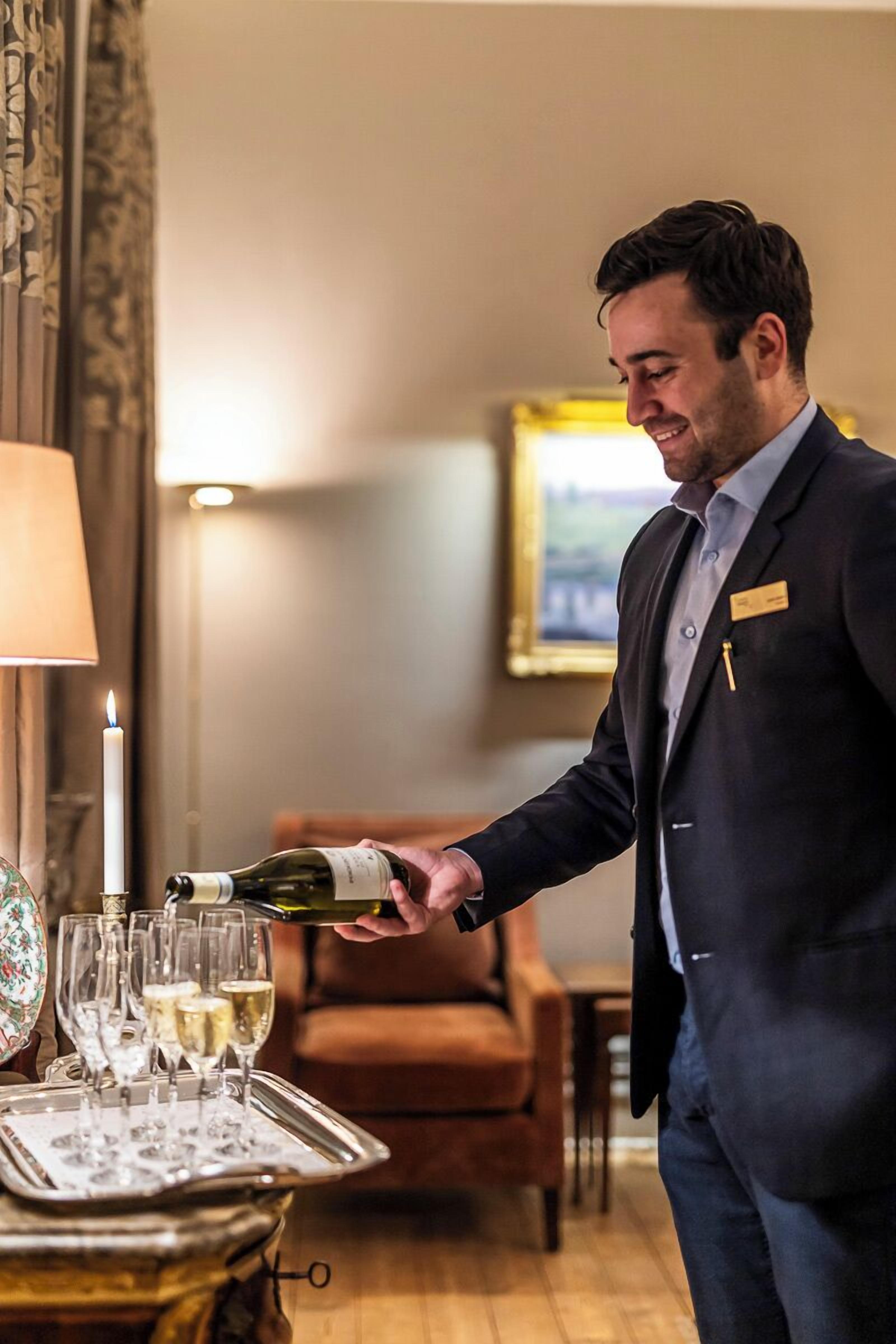 Een man schenkt champagne in glazen op een dienblad in een kamer met een lamp, een lijst met een foto en een bruine fauteuil.