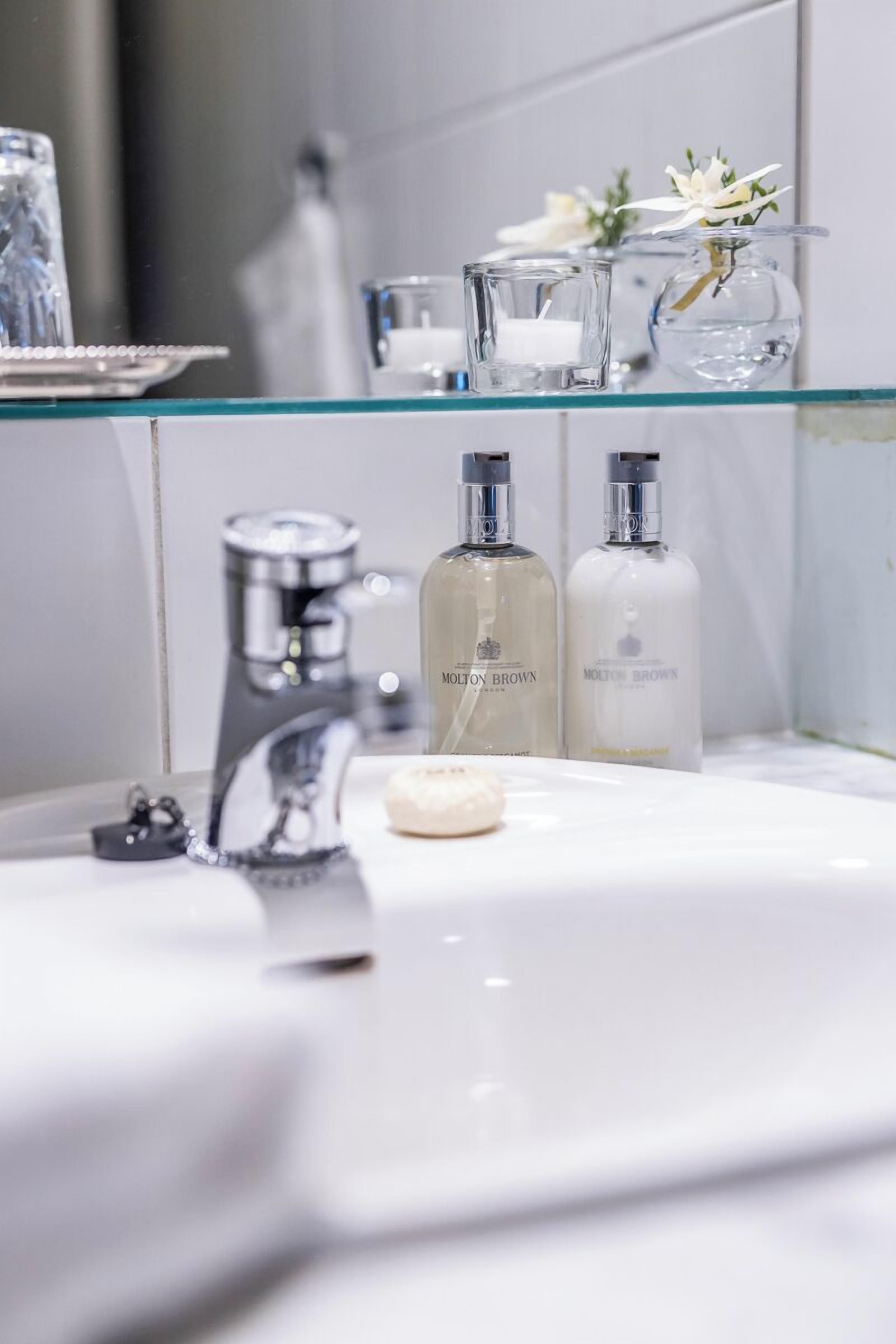 A bathroom with a glass shelf holding a candle, a glass, and a vase with flowers. There are two bottles of Molton Brown soap on the sink.