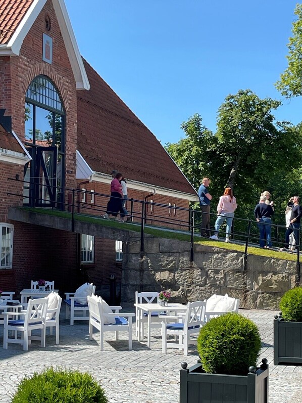 Ein Backsteinhaus mit geneigtem Dach, einer Außenbestuhlung mit weißen Stühlen und Tischen sowie einer Steinmauer.