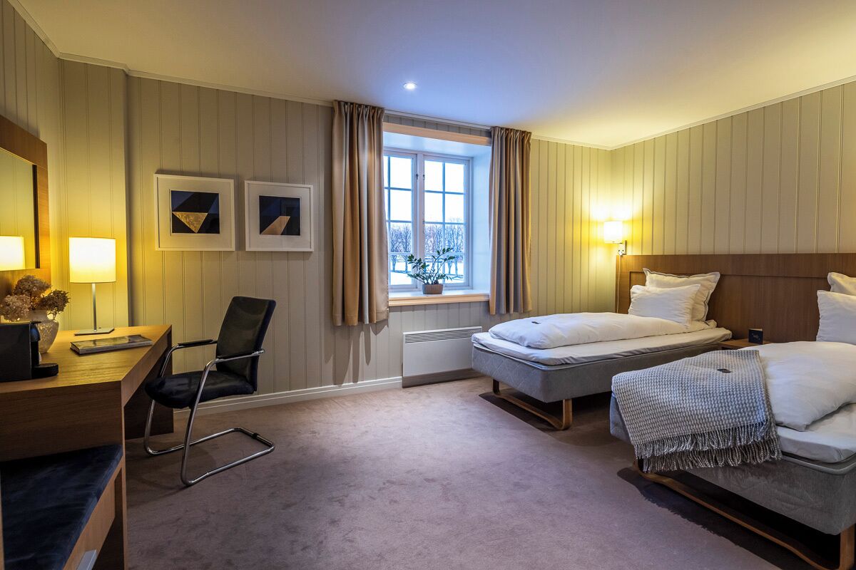 A hotel room with a desk, two beds, a chair, and a window with curtains.