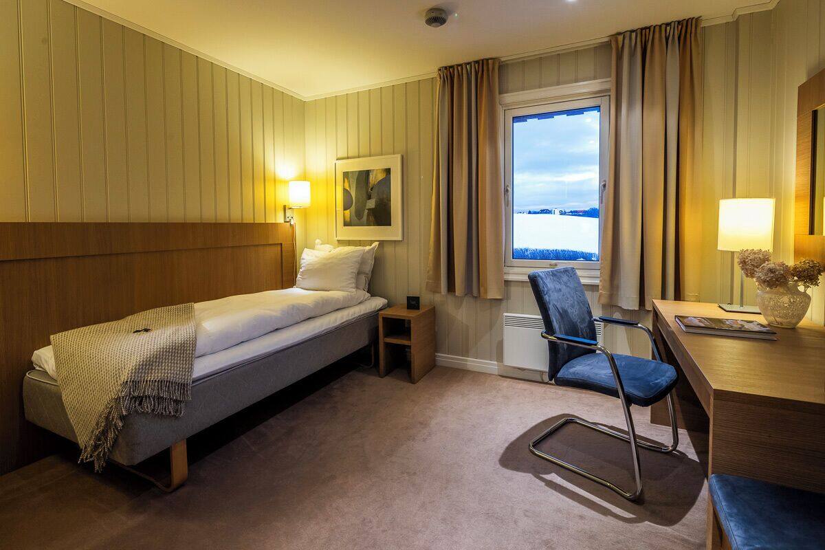 A hotel room with a bed, a desk, a chair, a window with curtains, and a lamp.