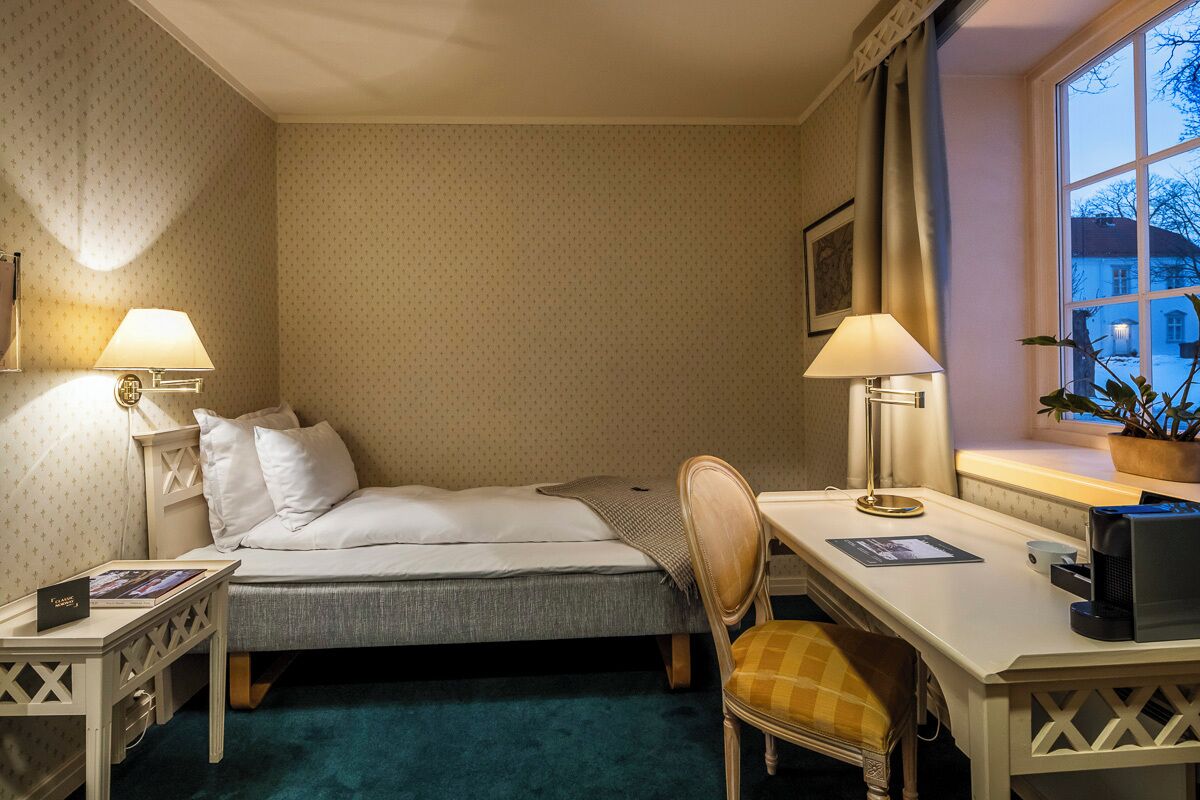 A hotel room with a bed, desk, chair, lamps, and a window with a view of buildings.