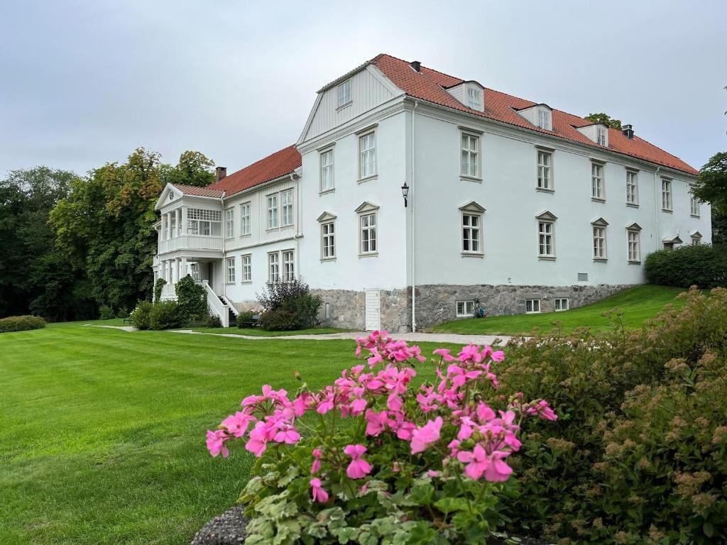 Un edificio grande y blanco con tejado rojo, rodeado de césped verde y flores rosadas.