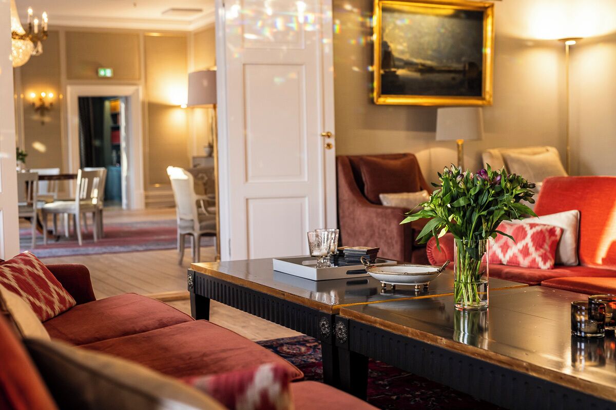 A hotel lobby with a red sofa, a coffee table, and a vase of flowers. There is a painting on the wall and a door leading to another room.