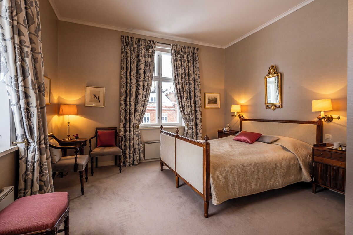 A bedroom with a four-poster bed, two bedside tables, a chair, a desk, and a window with curtains.