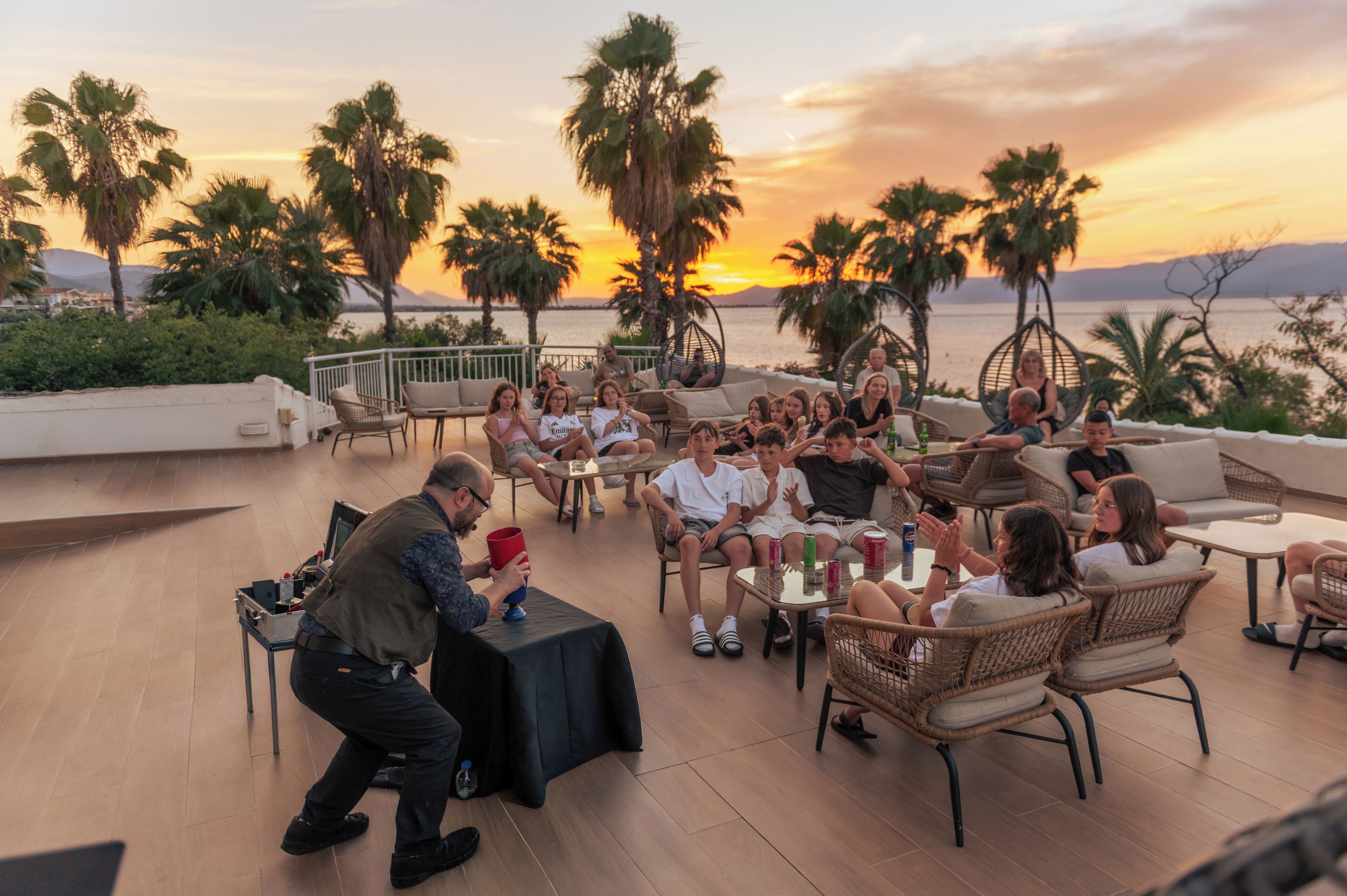 A group of people seated on outdoor furniture around a table, with a sunset view in the background.