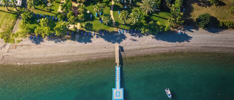 Vista aérea de una playa con un muelle de madera que se adentra en el agua, un pequeño bote cerca del muelle y una fila de sillas azules a lo largo de la orilla.