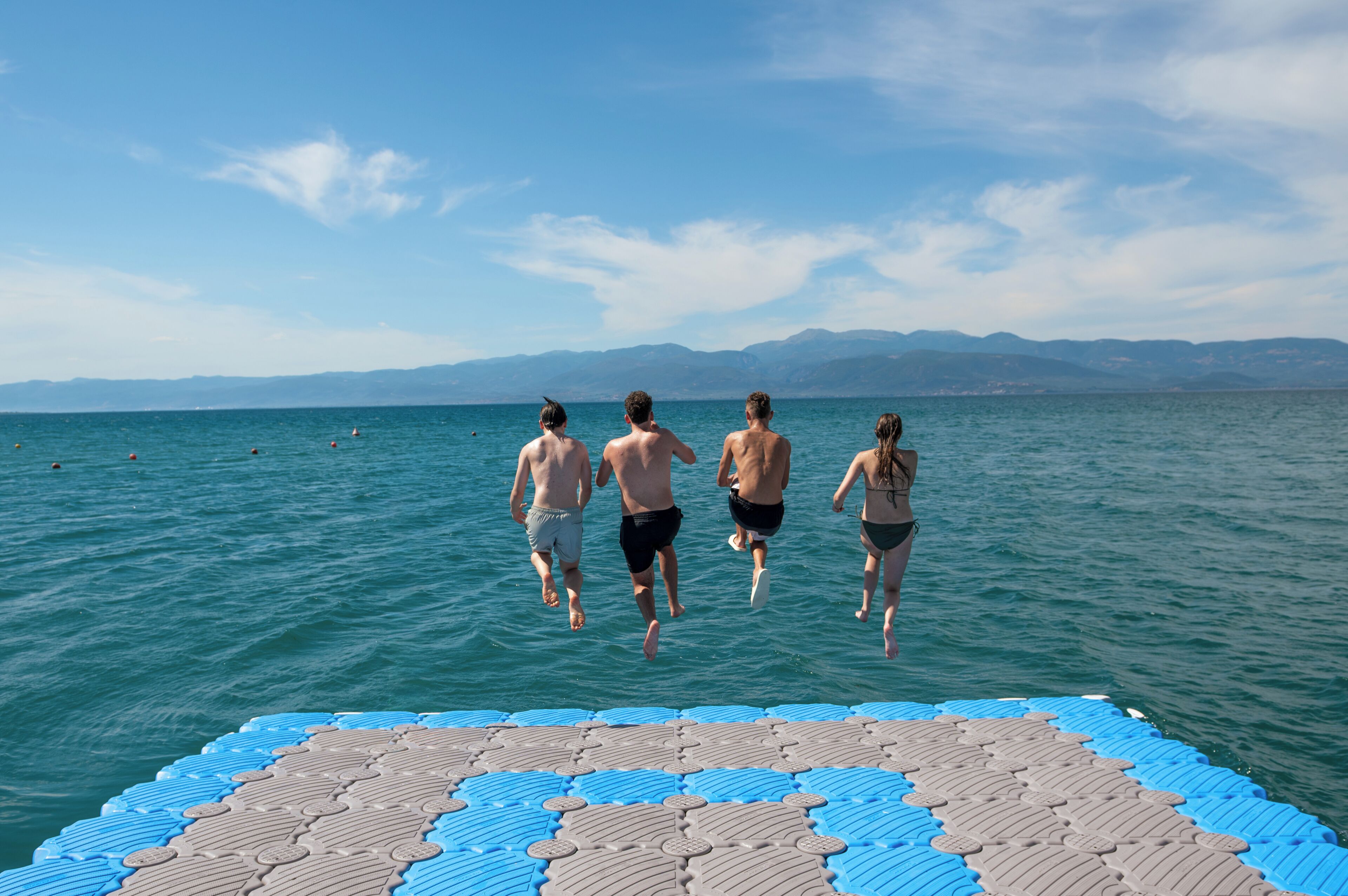 A group of people jumping off a blue and gray floating platform into a body of water with a mountainous landscape in the background.