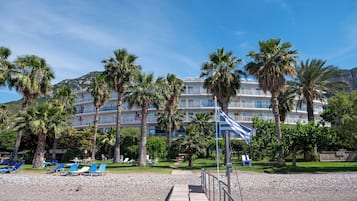 Hotel al costat de la platja amb nombroses balcons, palmeres i una platja de pedres amb gandules.