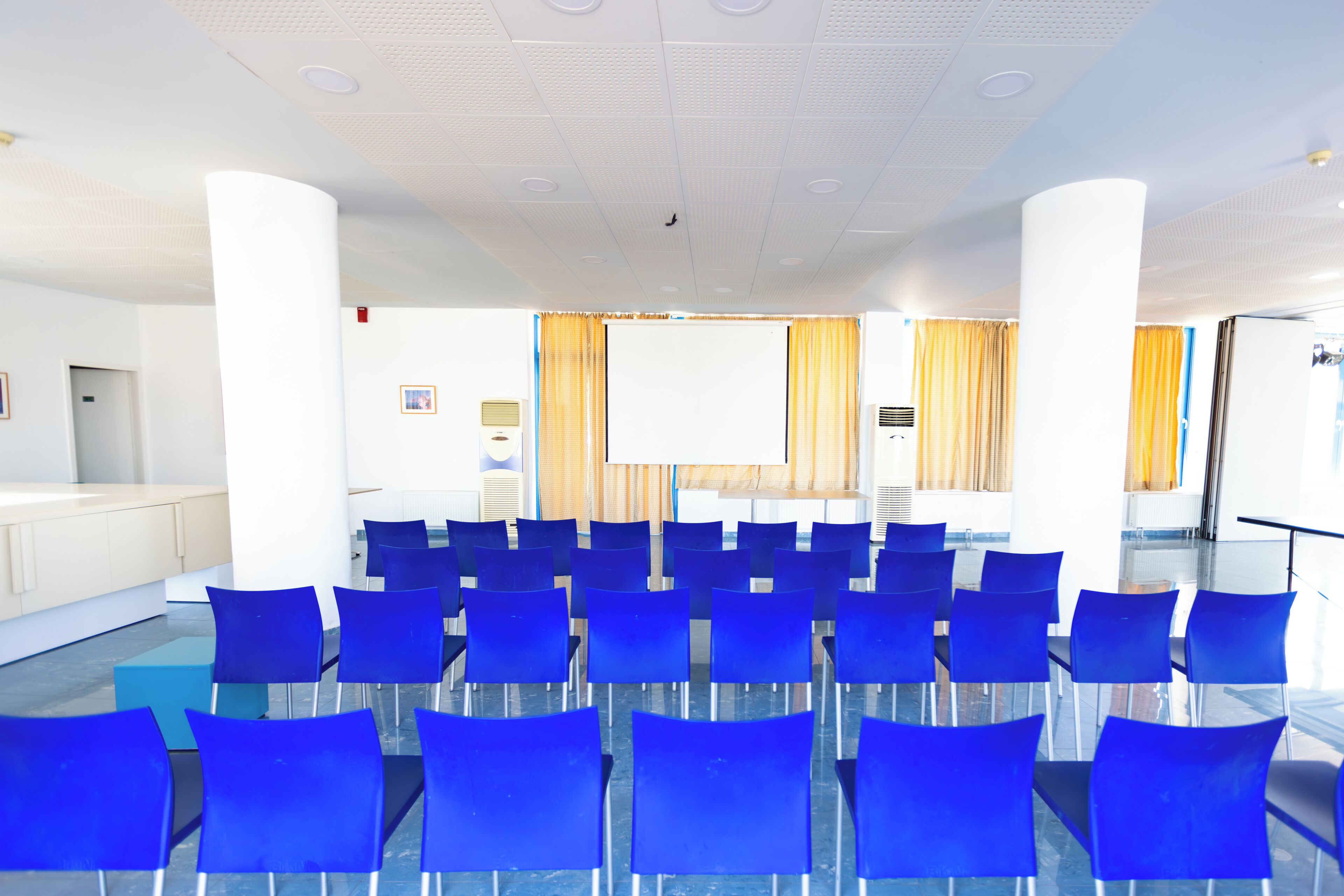 A conference room with rows of blue chairs, a large white screen, and a podium.