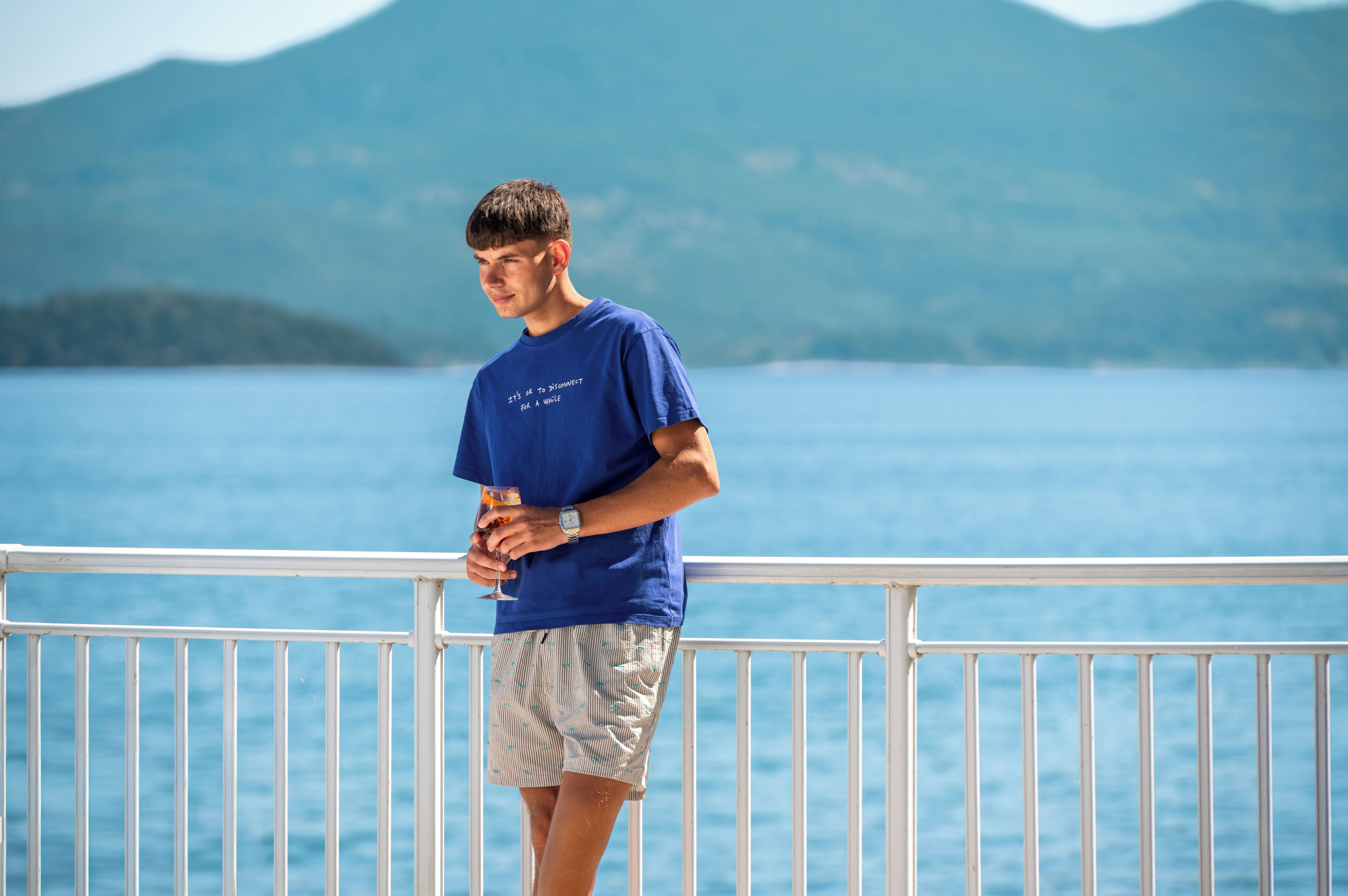 A person standing on a balcony with a railing, overlooking a body of water and a mountainous landscape.