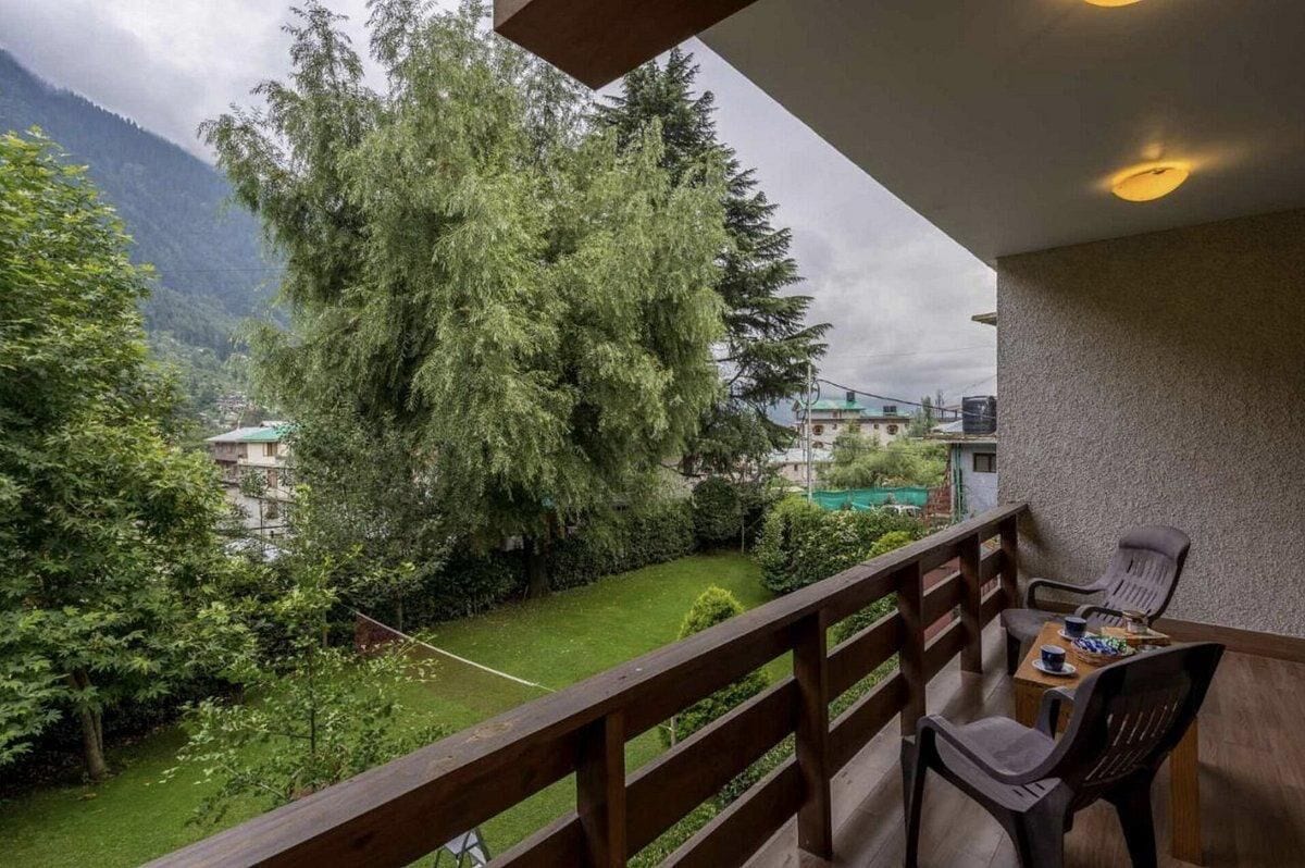 Un balcone con vista su un giardino rigoglioso e edifici in lontananza, ringhiera in legno e un tavolo con sedie.