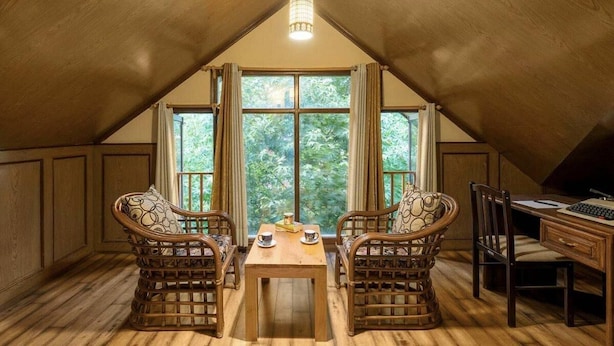 A cozy attic room with a wooden ceiling, two wicker chairs, a wooden desk, and a large window offering a view of greenery outside. - The Bear Creek Homestay Manali (Manali)