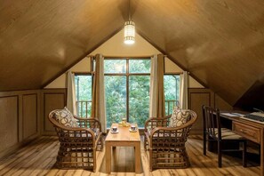 Una camera mansardata accogliente con soffitto in legno, due poltrone in vimini, una scrivania in legno e un'ampia finestra che si affaccia sul verde circostante.
