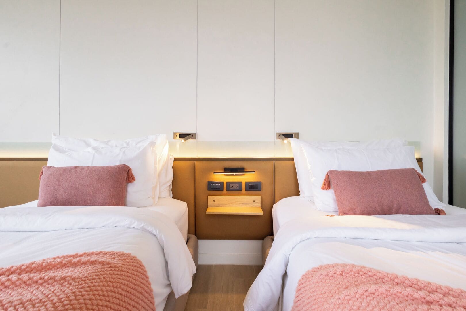 A hotel room with two beds, each with white bedding and pink throw pillows. There is a wooden nightstand between the beds with a small shelf and a wall-mounted control panel.