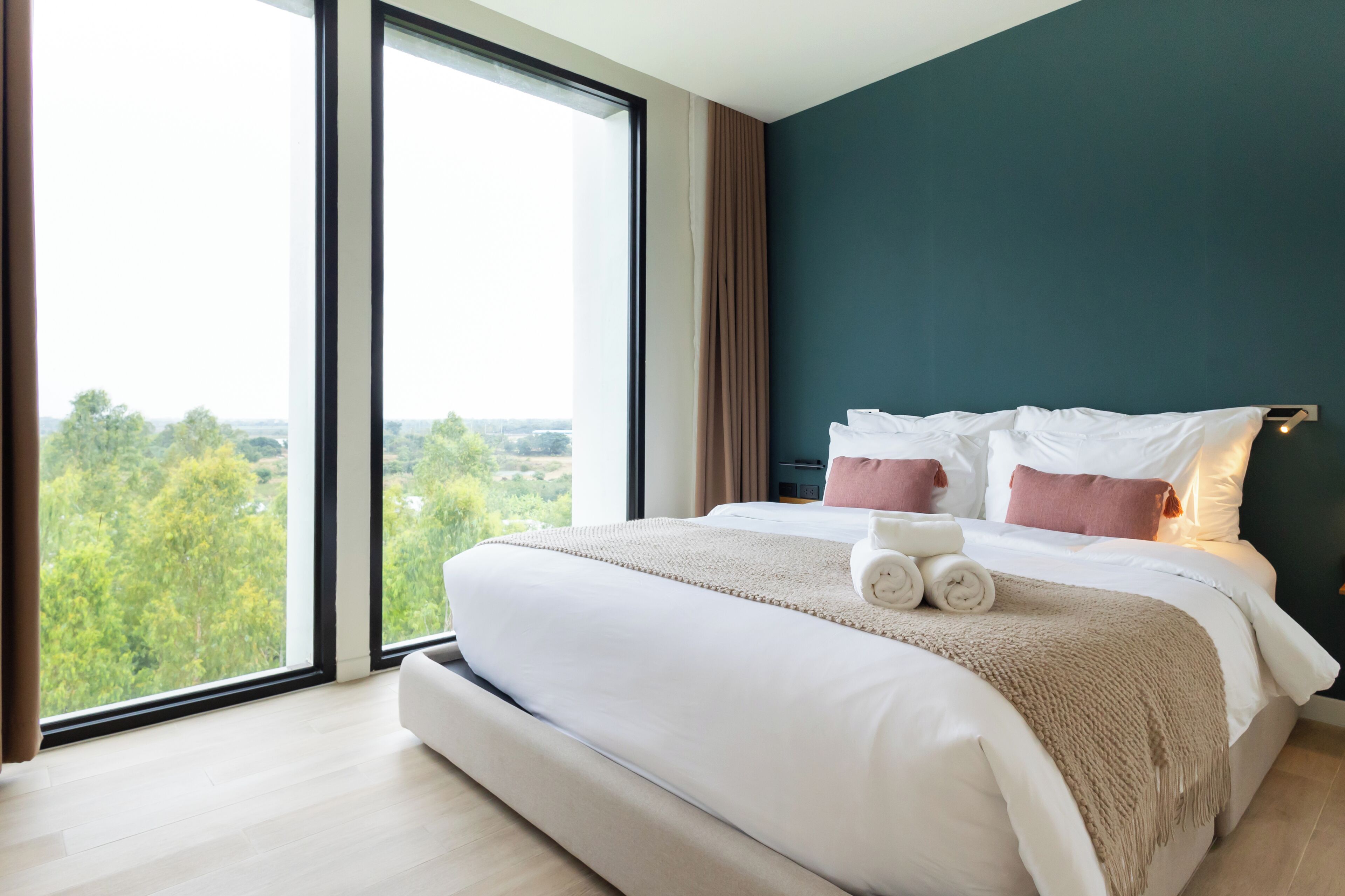 A modern hotel room with a large bed, a view through floor-to-ceiling windows, and a teal accent wall.