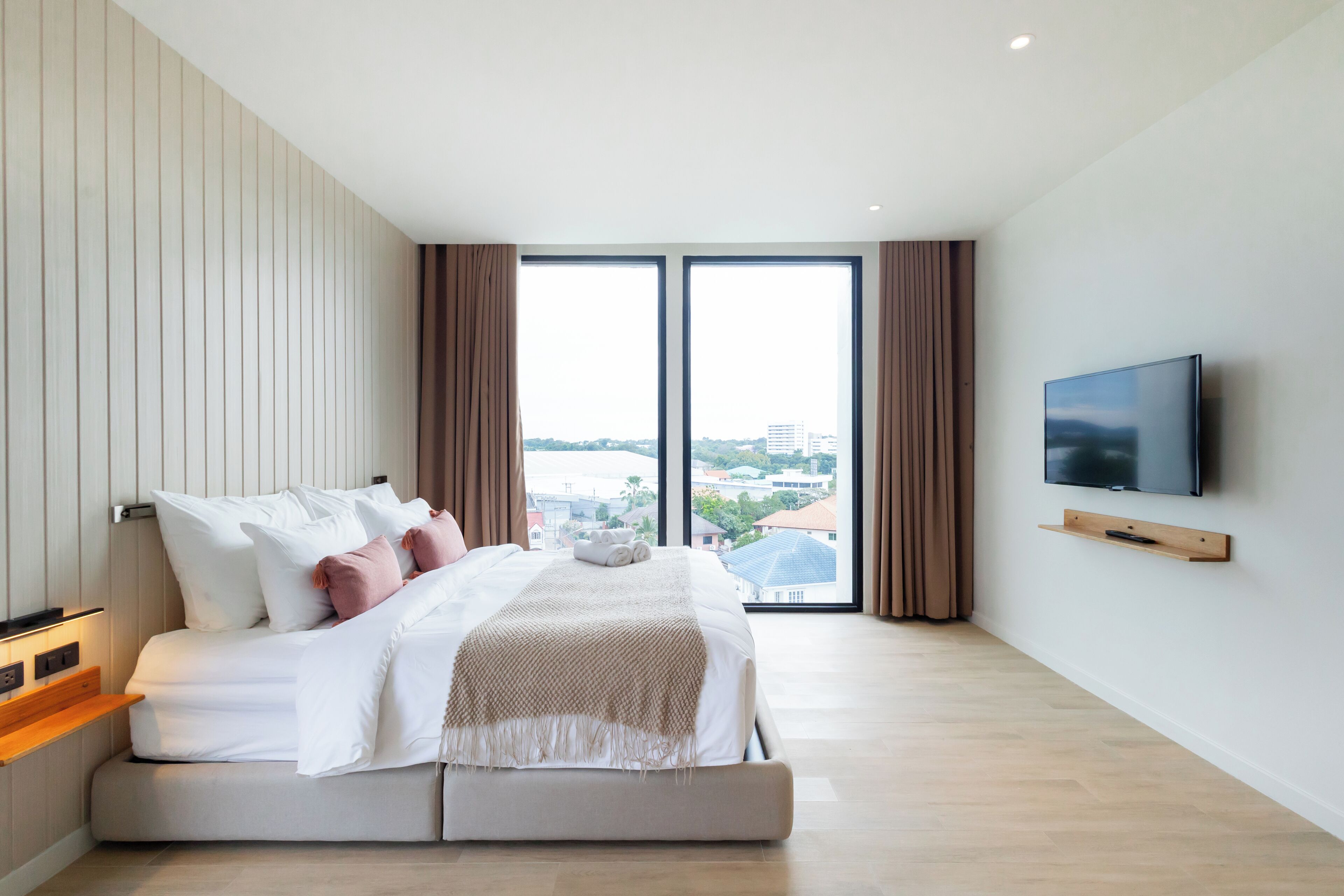 Una habitación de hotel moderna con una cama grande, un televisor de pantalla plana y vistas a la ciudad a través de ventanas de gran altura.
