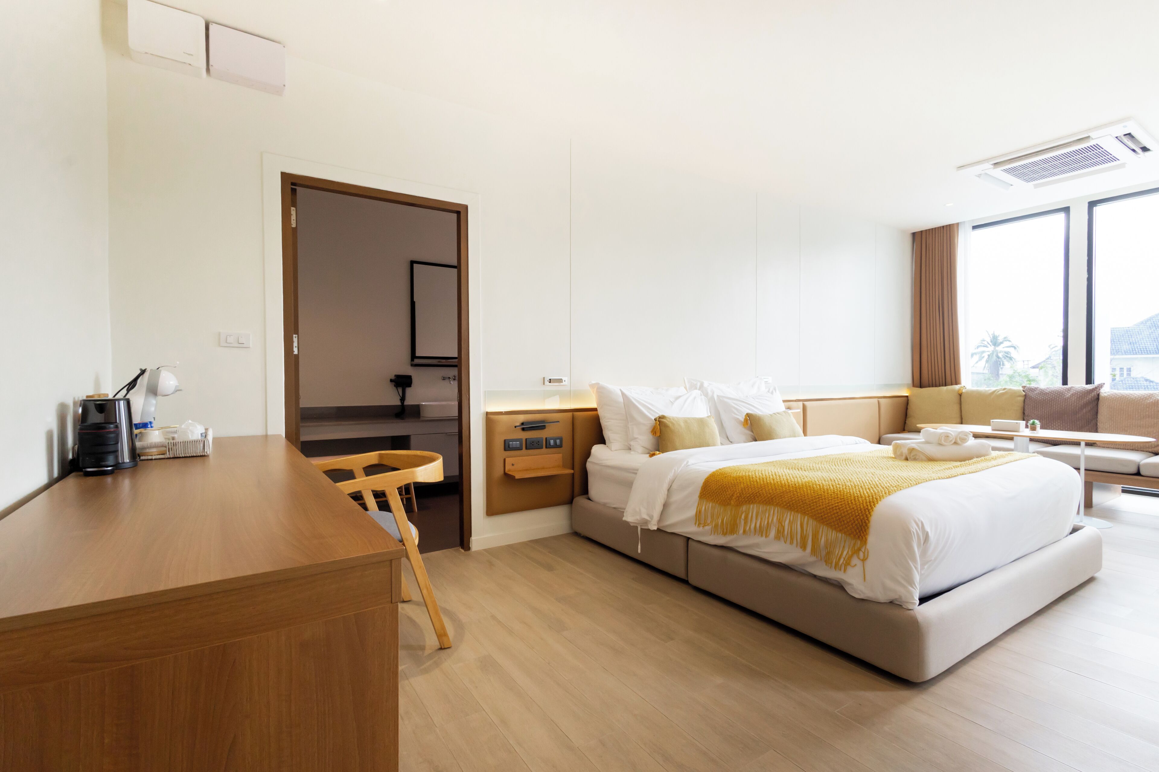 A modern hotel room with a wooden desk, a bed with a yellow throw, and a large window offering a view.