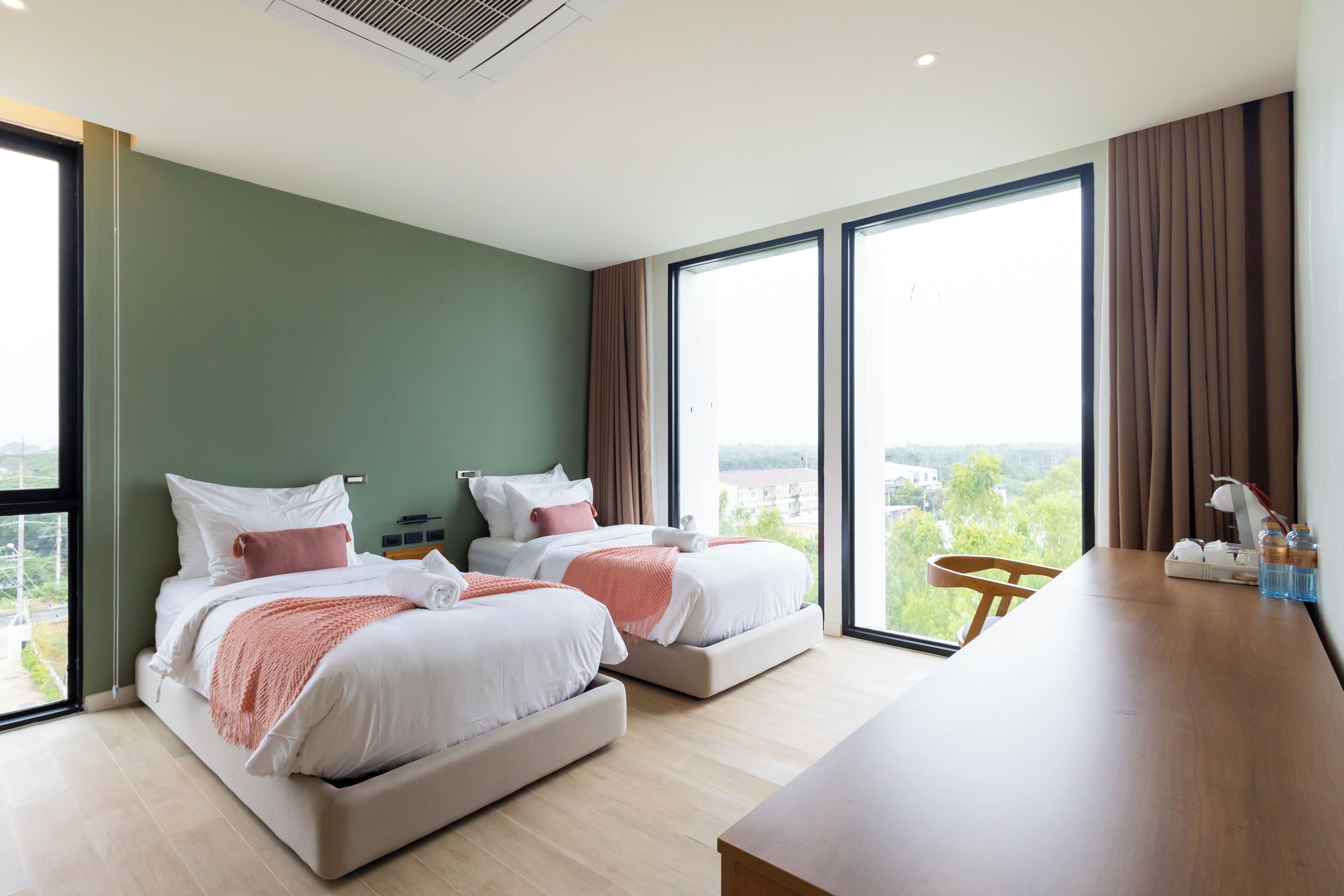 A modern hotel room with two beds, a wooden desk, and large windows offering a city view.