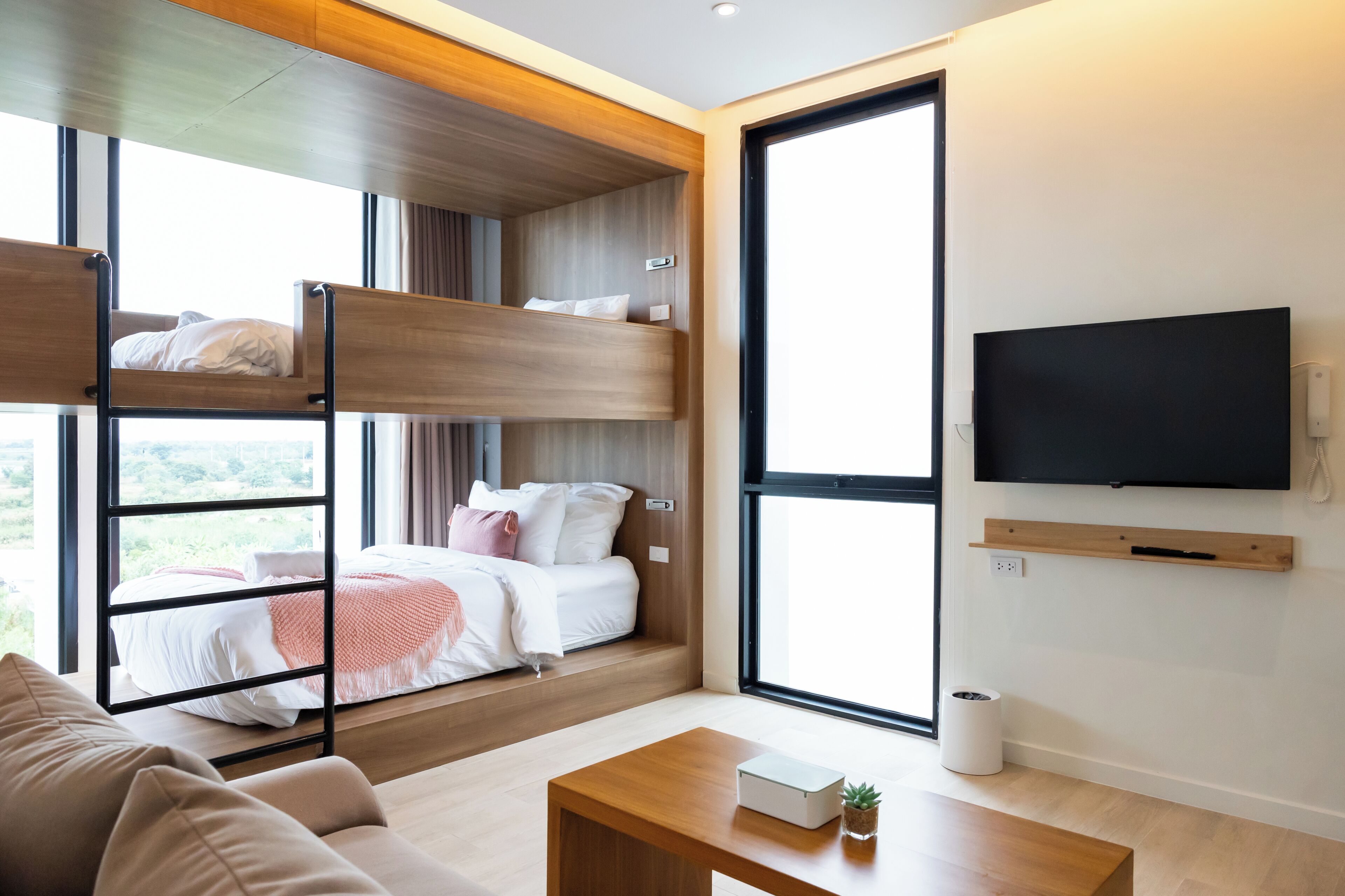 A modern hotel room with a bunk bed, a flat-screen TV mounted on the wall, a wooden coffee table, and a large window offering a view of the outdoors.