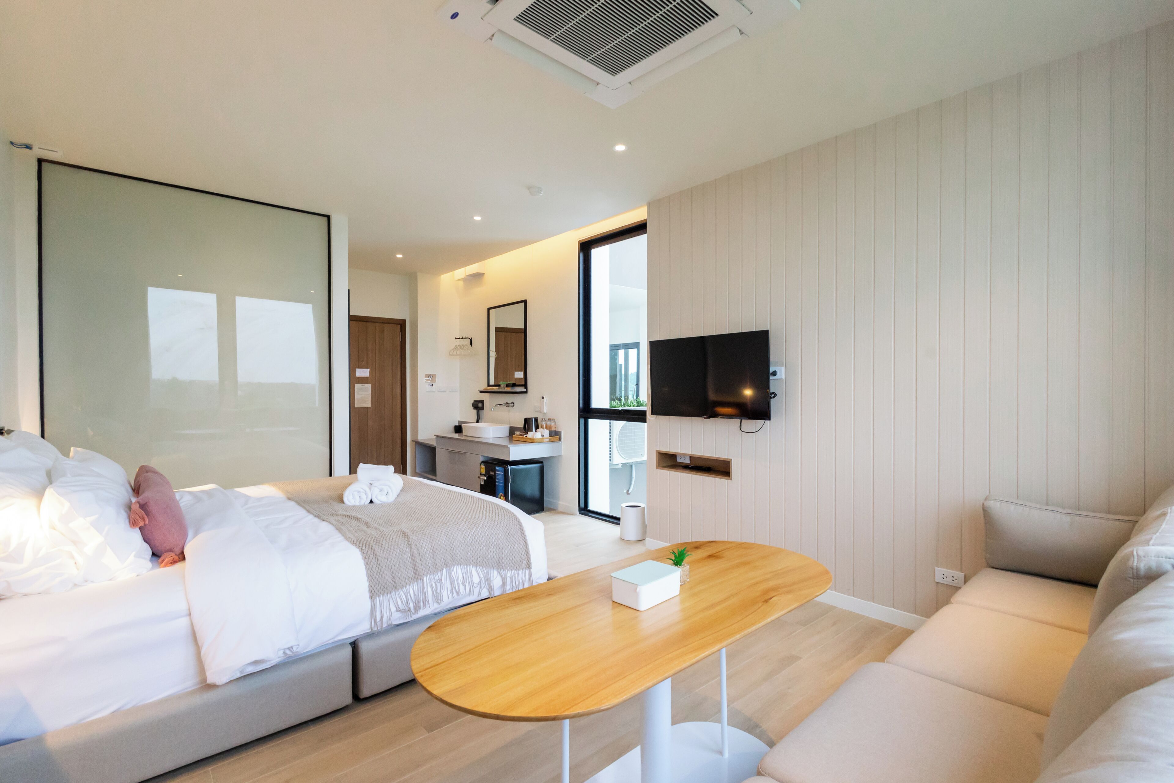 Une chambre d’hôtel moderne, avec un lit, un canapé, une table en bois, une télévision et une salle de bain visible au fond.