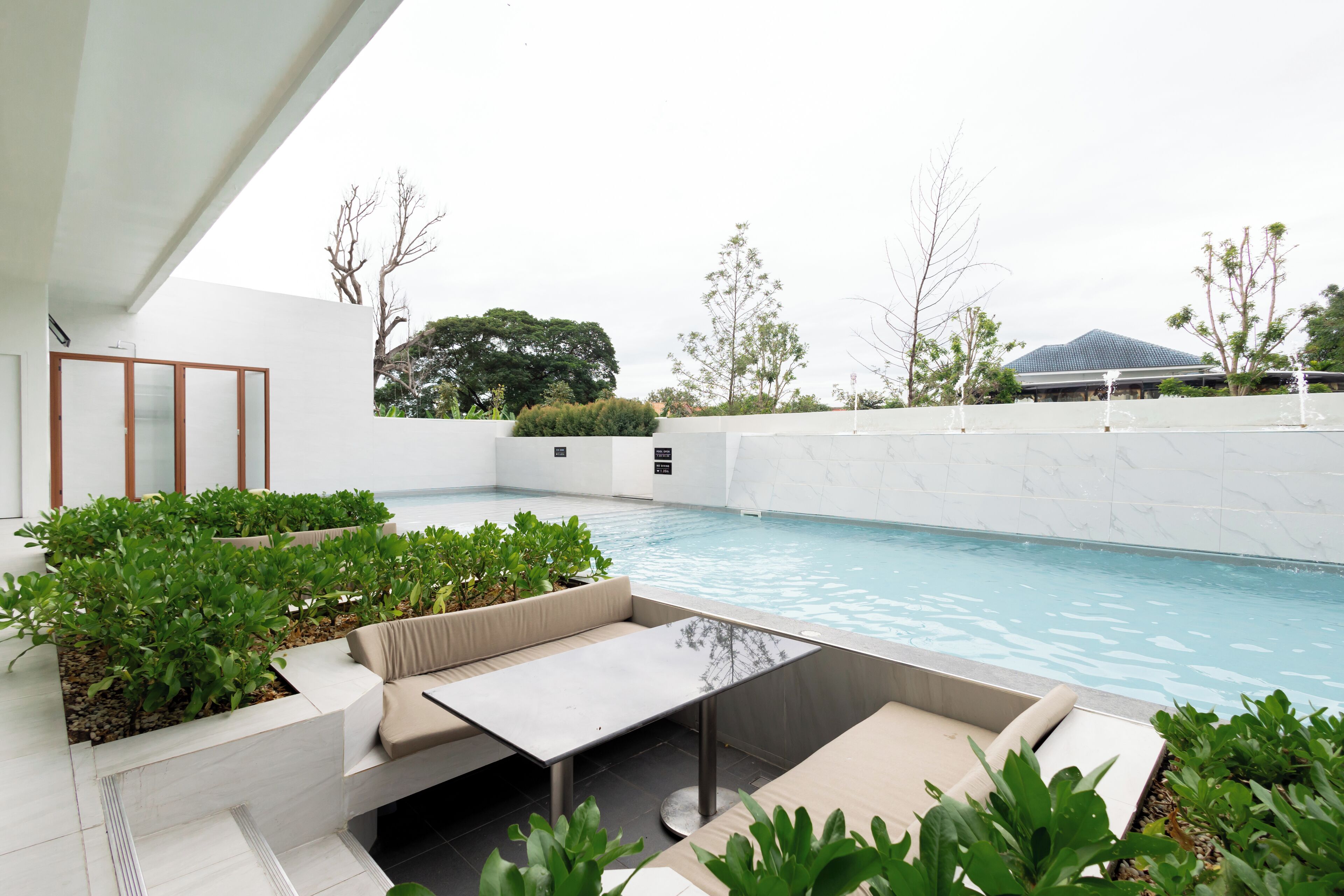 A modern outdoor pool area with a seating area, plants, and a view of a building in the background.