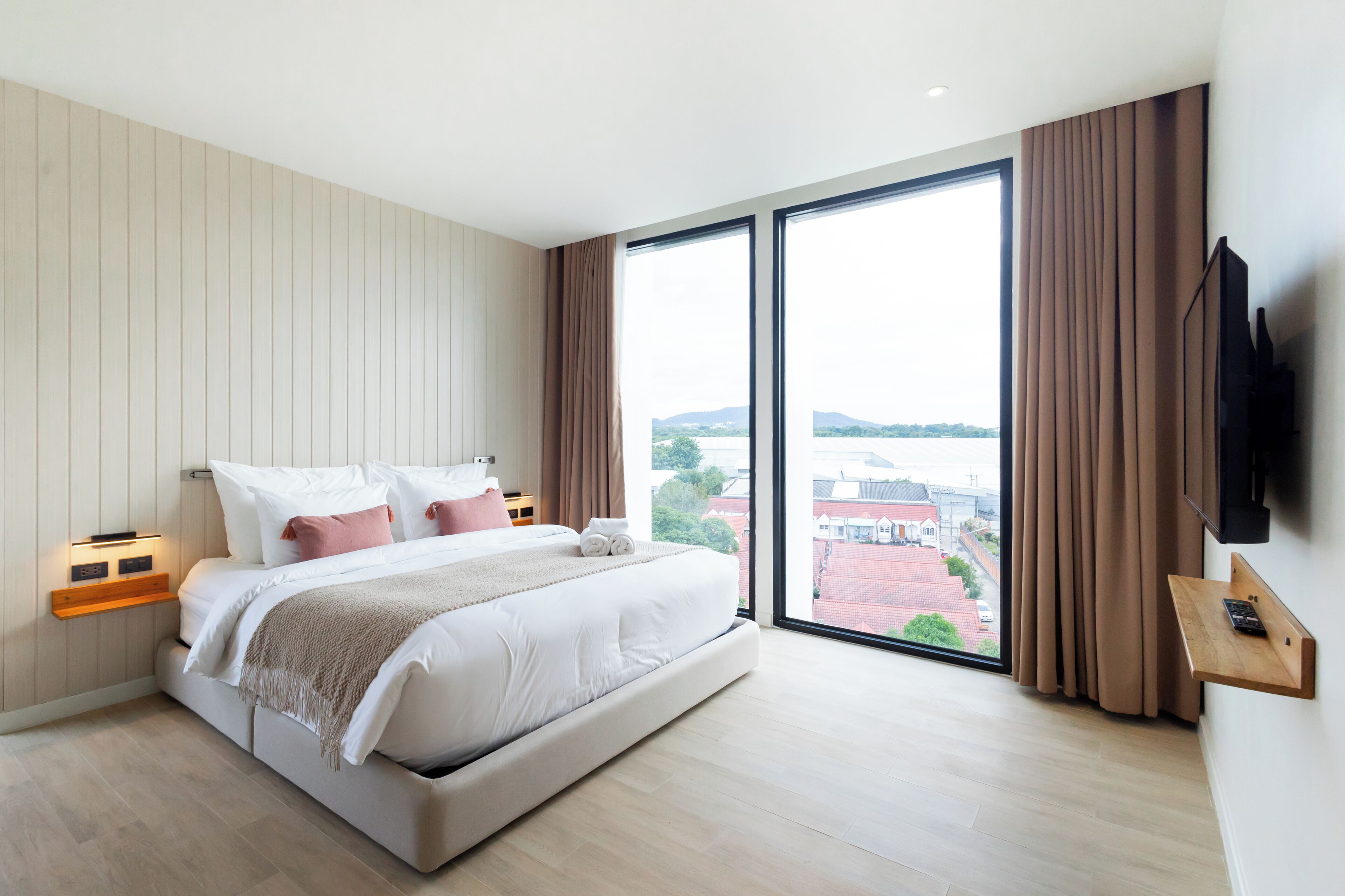 Un dormitorio moderno con una cama grande, un televisor montado en la pared y vistas de la ciudad a través de amplias ventanas.