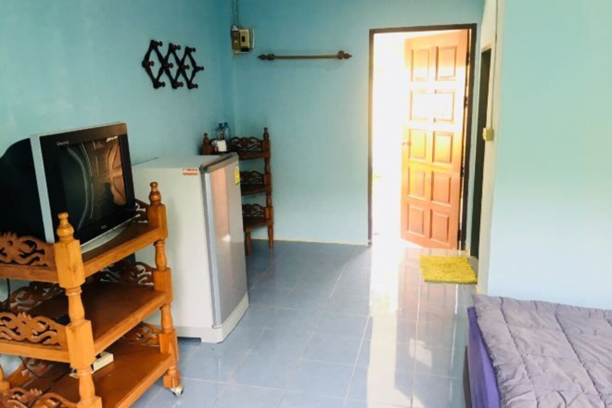 A room with a bed, a TV on a wooden stand, a refrigerator, and a door leading to another room.