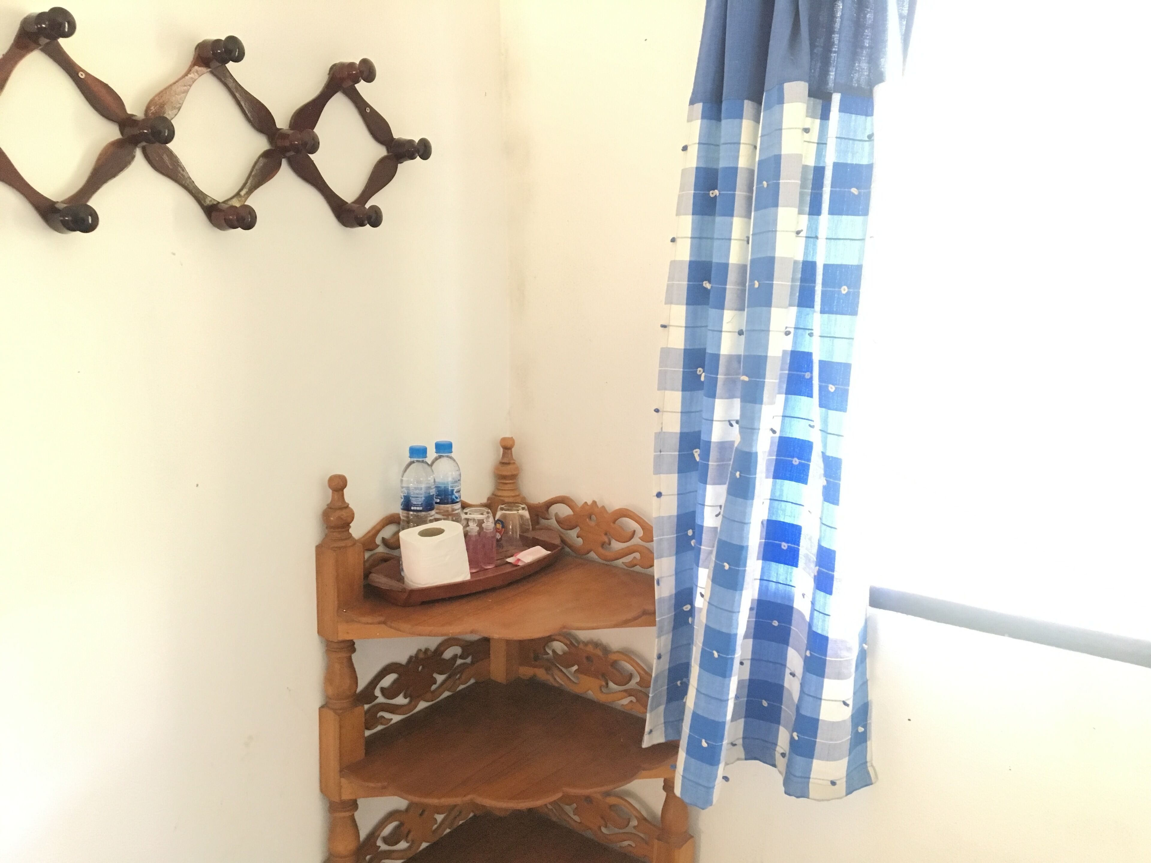 Rincón de una habitación con un estante de madera que contiene artículos de aseo, un adorno de pared y una cortina a cuadros.