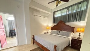 Una camera da letto con un letto di legno, due comodini con lampade, una ventola a soffitto, una finestra con tende e una porta che conduce a un'altra stanza.
