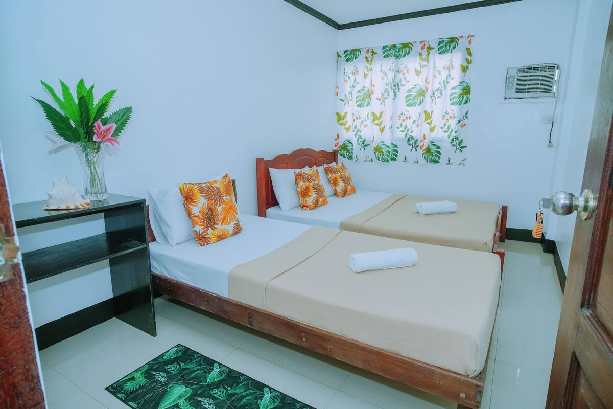 A hotel room with two beds, a nightstand, a vase with flowers, and a window with a tropical curtain.