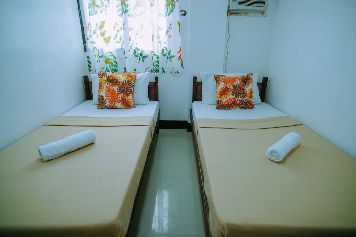 Two single beds with beige linens, each with a white pillow and an orange pillow with a leaf pattern.