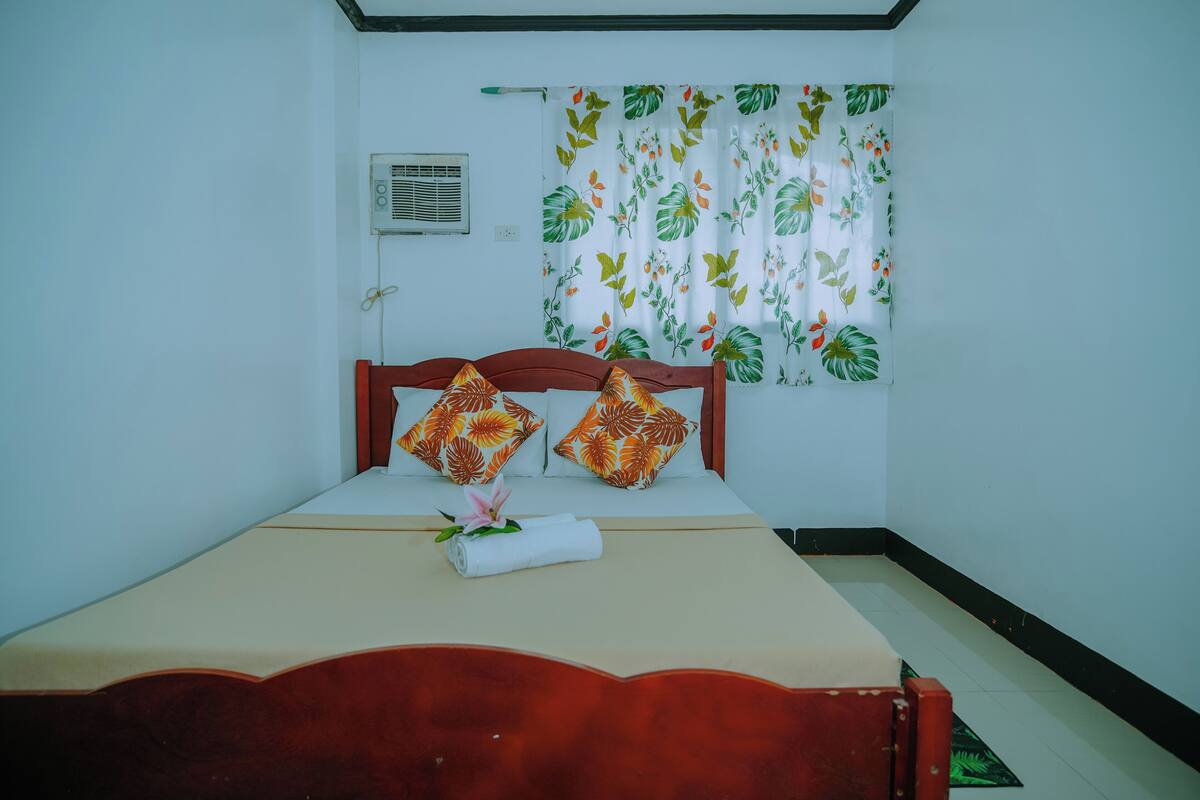 A neatly made bed with two pillows, a rolled towel, and a flower on the bed.