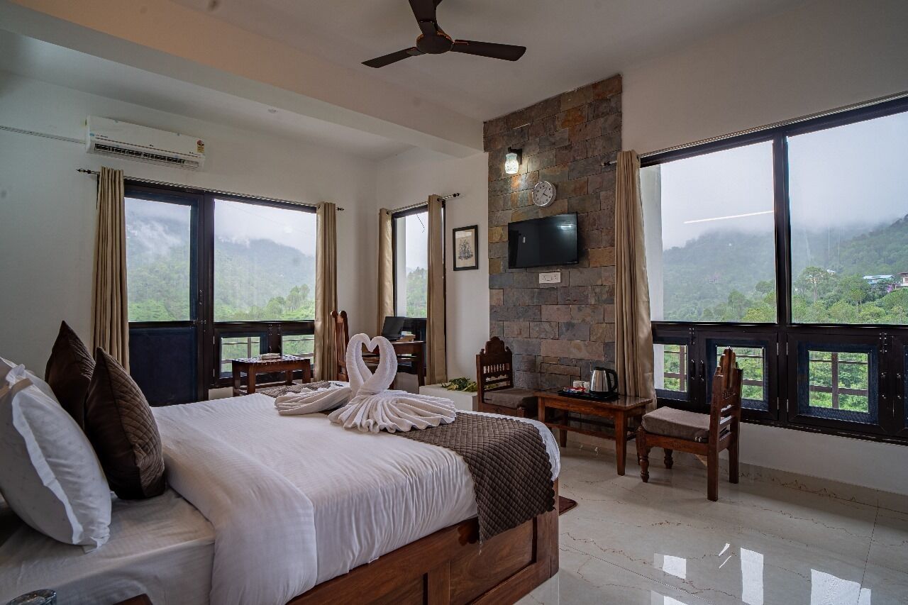 Habitación de hotel con cama, escritorio, silla, televisor y ventanas amplias con vistas a la montaña.