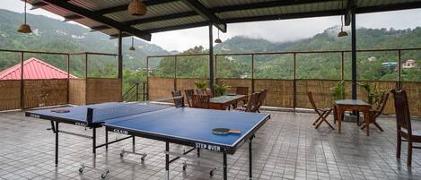 A ping pong table set up on a tiled patio with chairs and tables, overlooking a mountainous landscape.