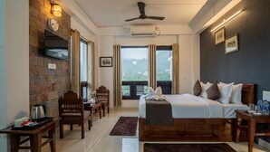 A hotel room with a large bed, a wooden headboard, a flat-screen TV mounted on a stone wall, a ceiling fan, and a view of greenery through the windows. - Captain Resort (Kasauli)