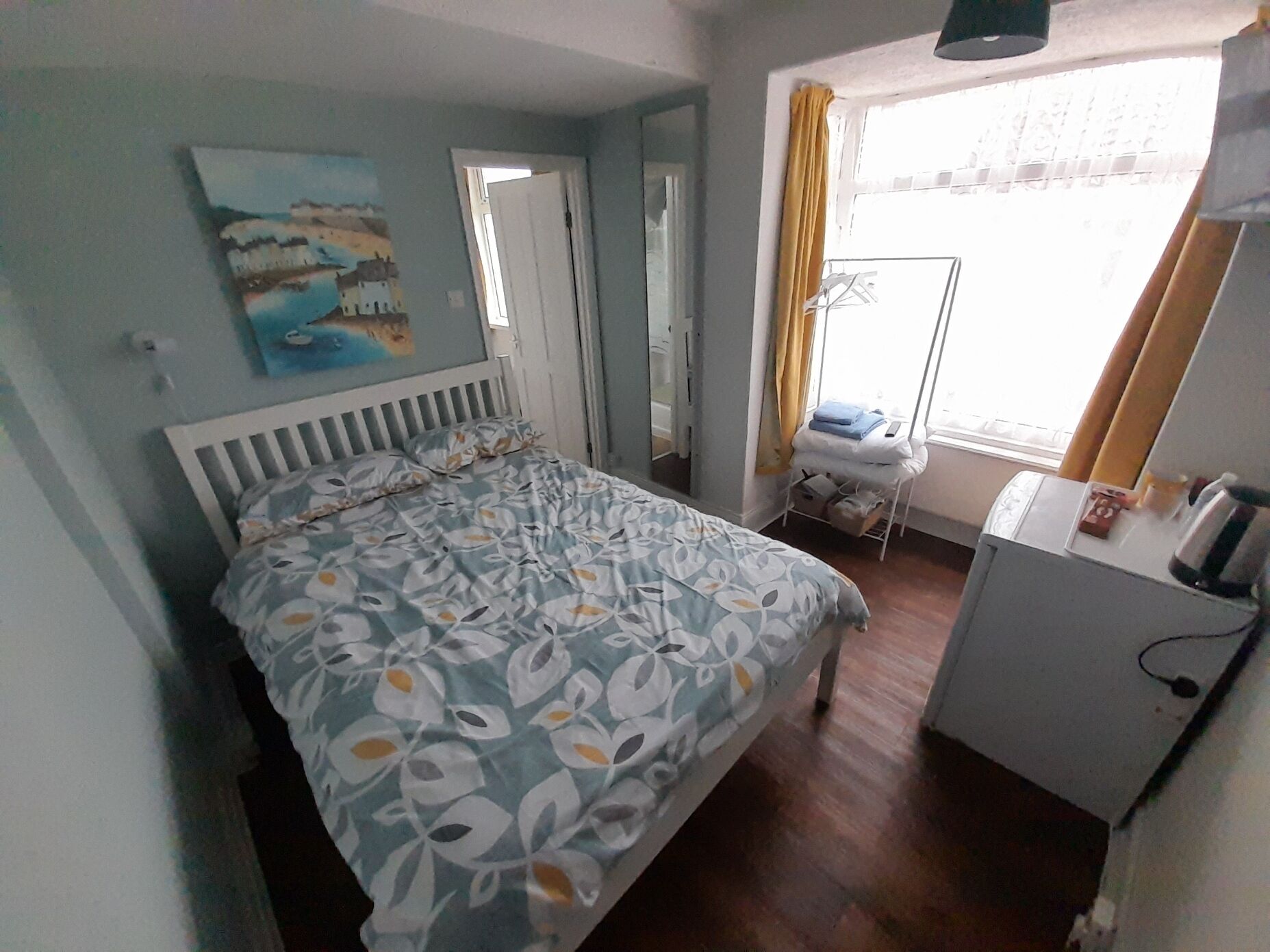 A bedroom with a bed, a bedside table, a wardrobe, a window with curtains, and a painting on the wall.