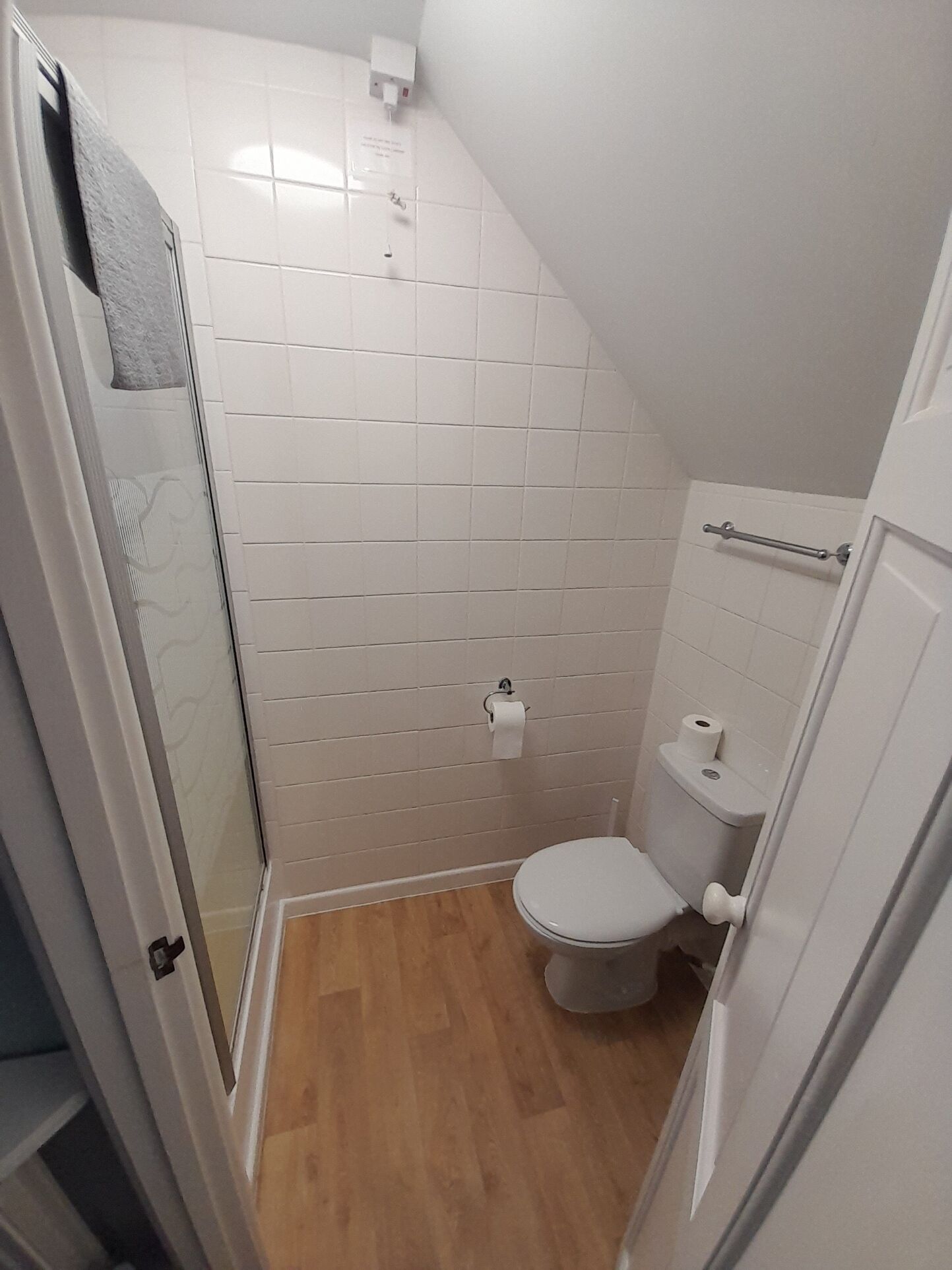 A small, white-walled bathroom with a toilet, a shower area, and a wooden floor.