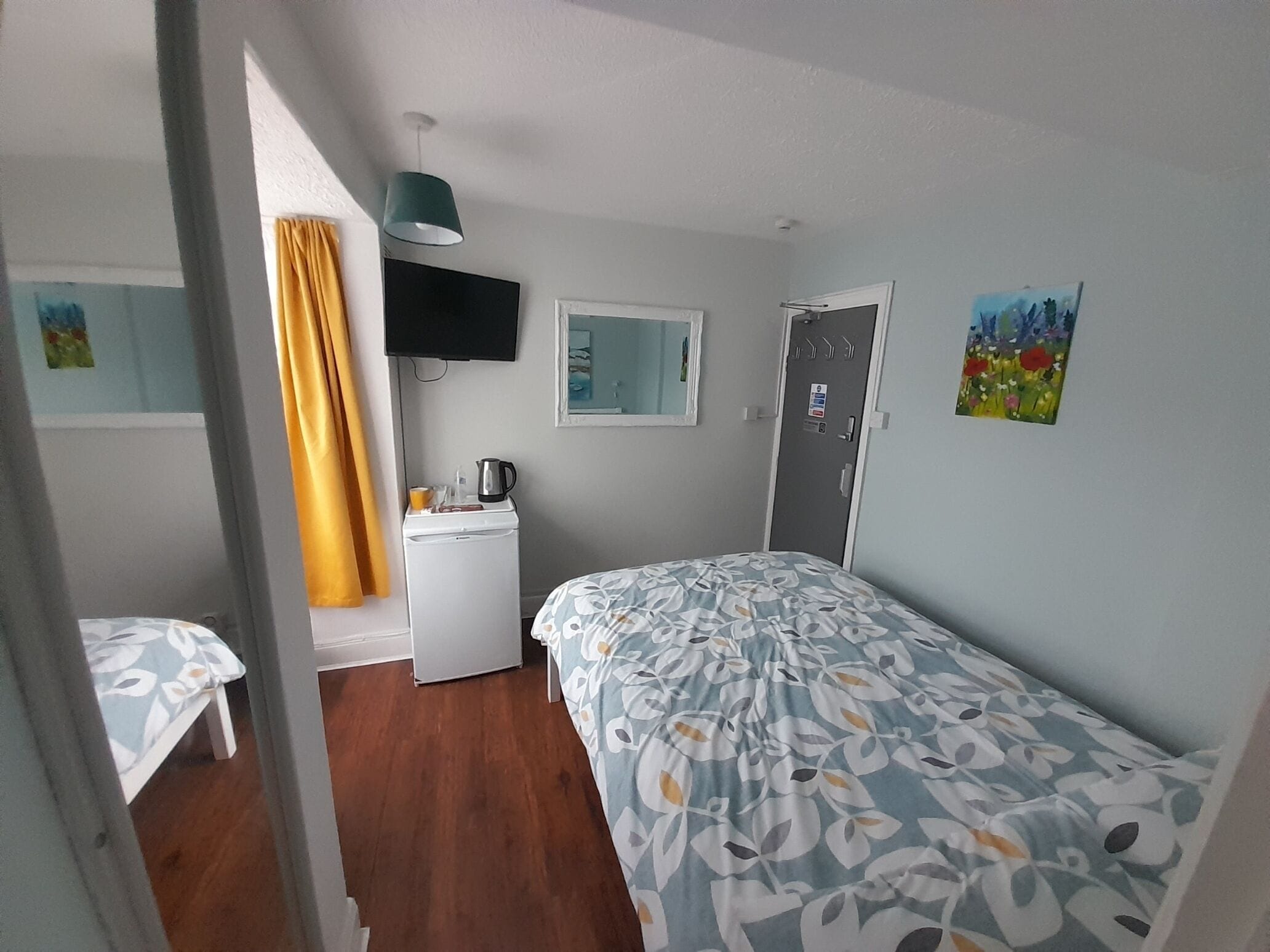 A small, well-lit room with a bed, a desk, a television, and a door.