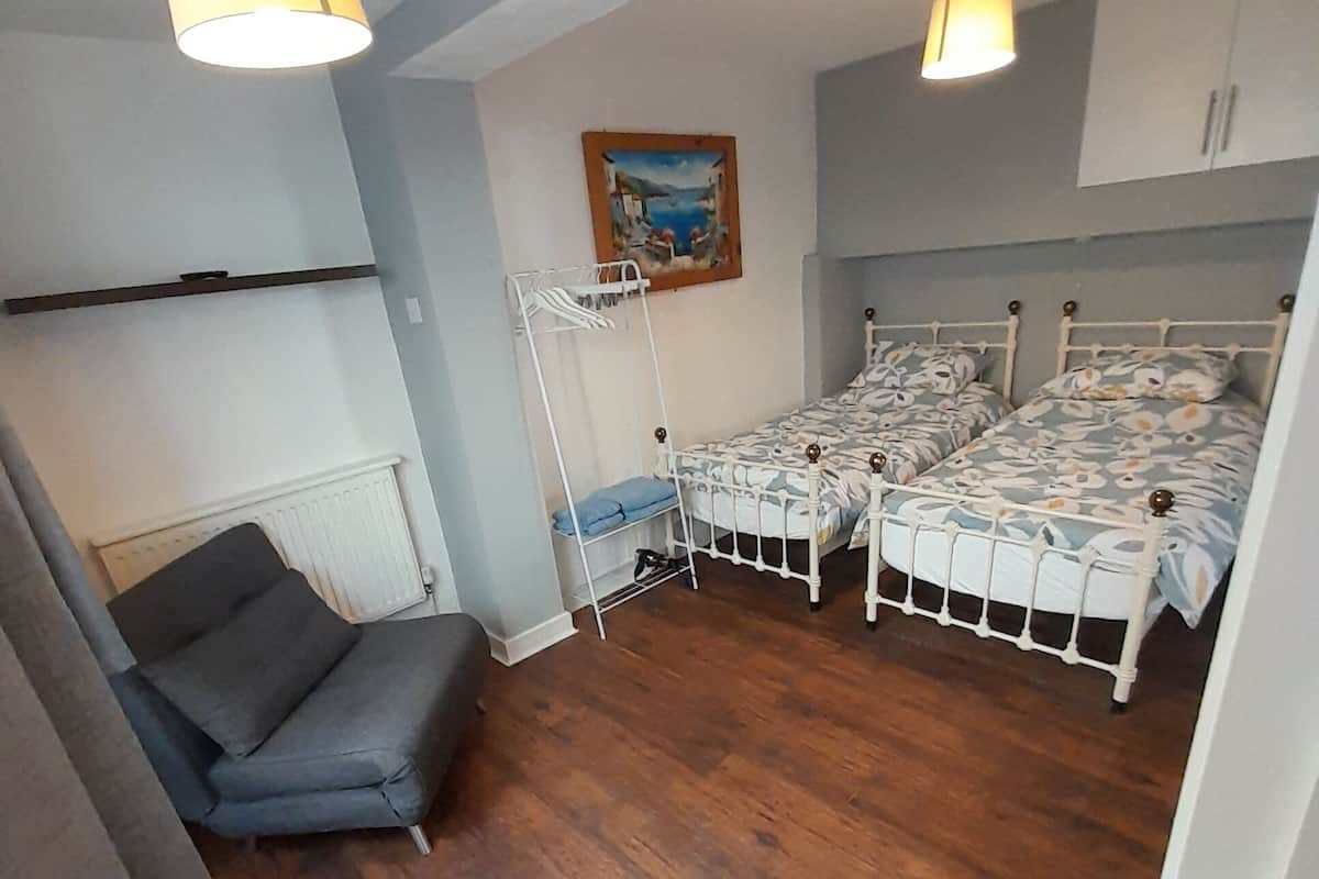 A bedroom with two beds, a chair, a radiator, and a painting on the wall.