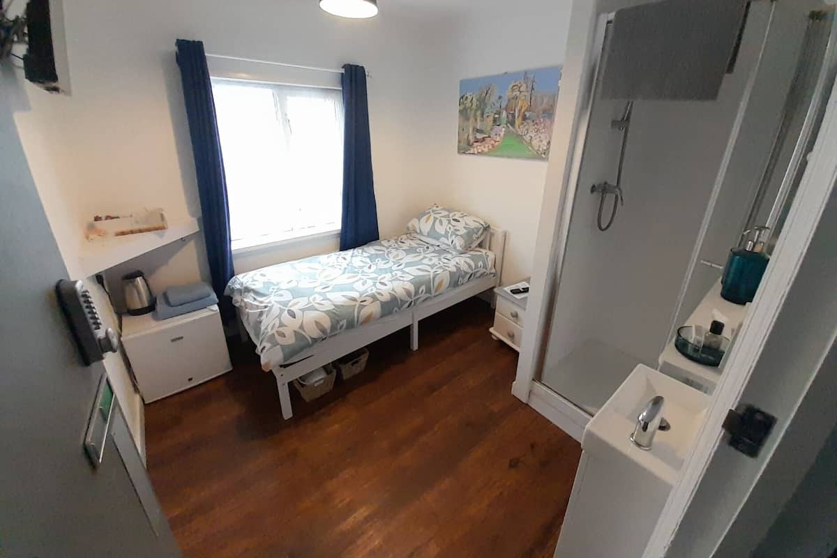 A small, compact bedroom with a single bed, a bedside table, a nightstand, a window with curtains, a painting on the wall, and a shower area.
