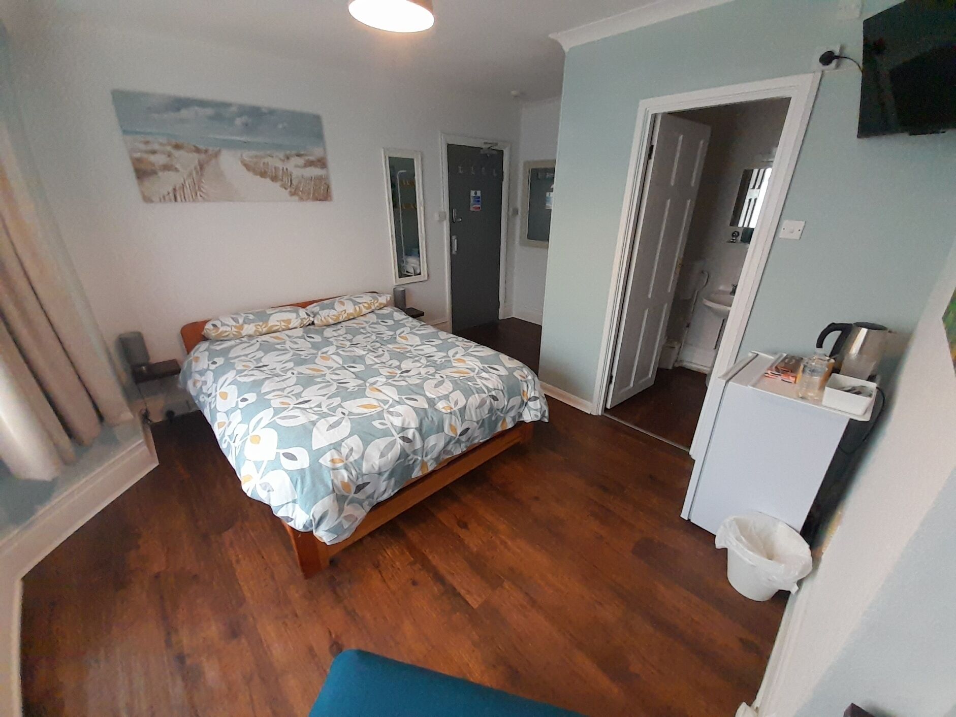Quarto com cama, soalho de madeira, banheiro com chuveiro e uma pequena cozinha.