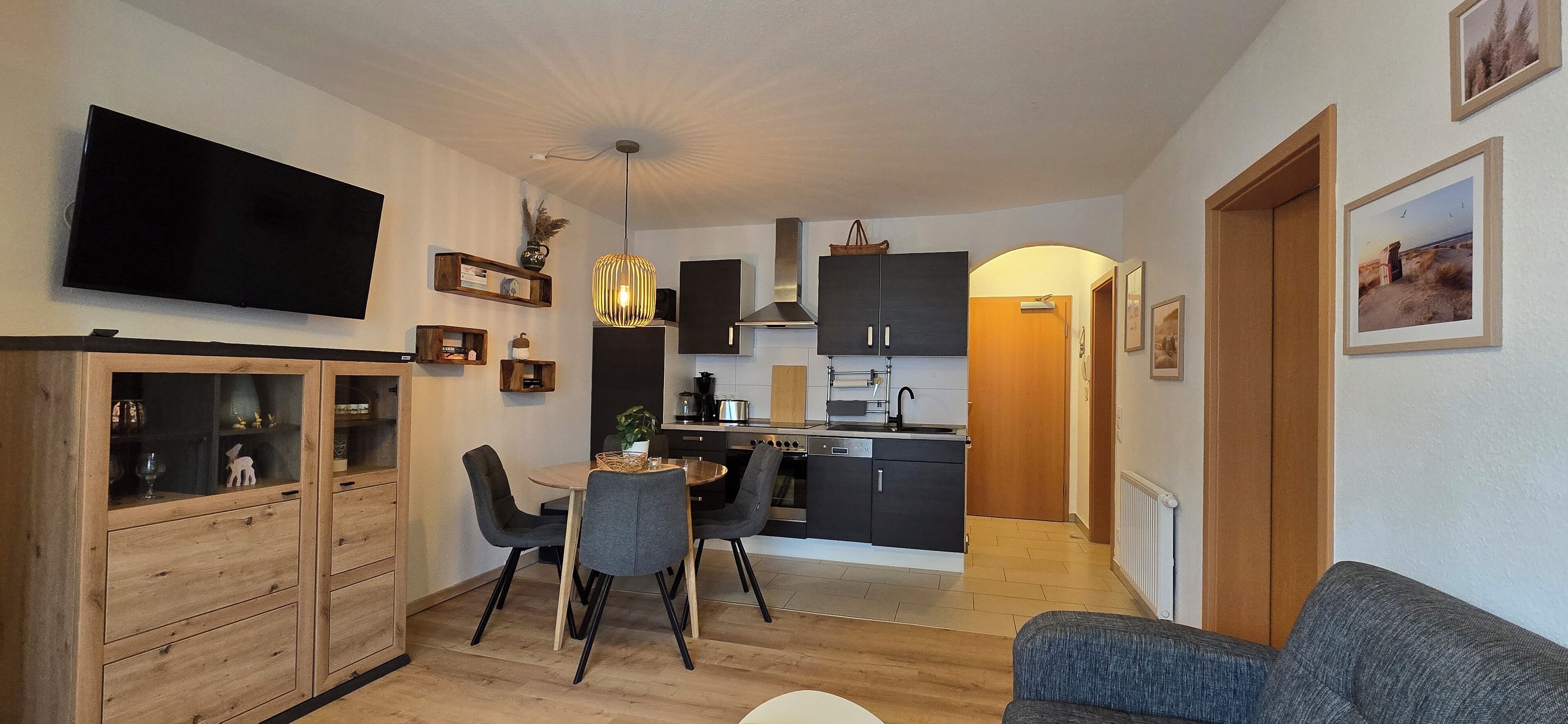 Una cocina moderna con armarios oscuros, un comedor con una mesa redonda y sillas, un televisor en la pared y una zona de estar acogedora con un sofá.