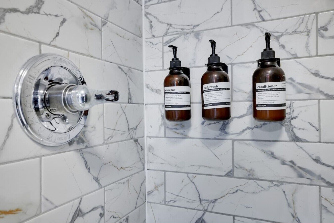 A bathroom with a marble wall, a chrome faucet, and three bottles of toiletries.
