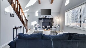 A modern living room with a dark sofa, a fireplace, and a staircase with wooden beams. - The Cozy Modern Cabin – Ski & Relax (Tobyhanna)