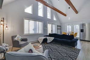 Un salon spacieux avec un plafond haut, de grandes fenêtres, un canapé bleu foncé, un fauteuil gris et un tapis à motifs.