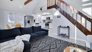 A modern living room with a dark sofa, a patterned rug, a staircase with wooden railings, and a kitchen in the background. - The Cozy Modern Cabin – Ski & Relax (Tobyhanna)