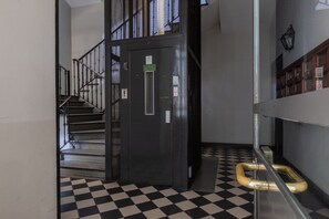 Ein Flur mit schwarz-weiß kariertem Boden, einem Metallaufzug und einer Treppe mit Metallgeländern.