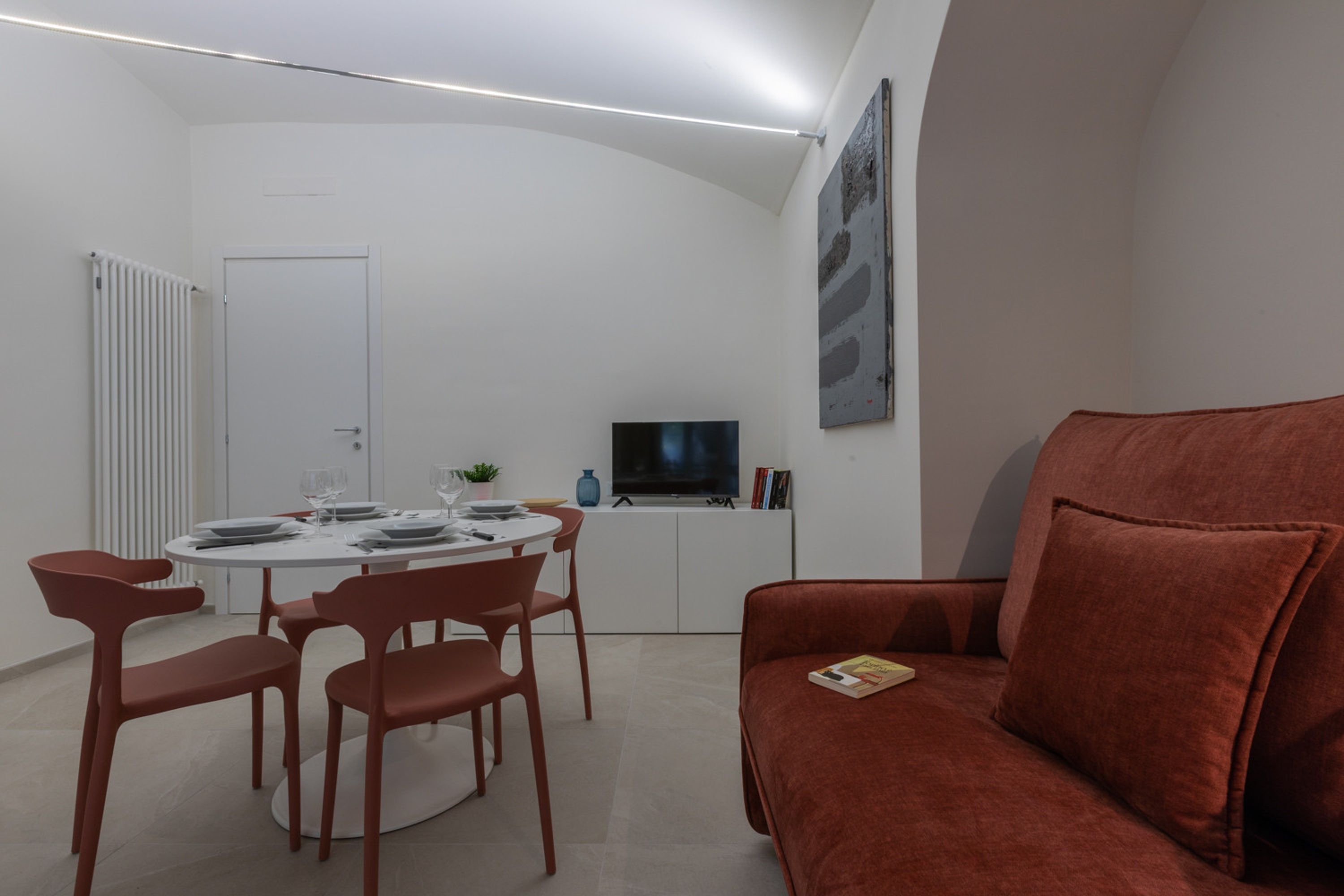 Ein modernes Wohnzimmer mit einem roten Sofa, einem Essbereich mit rundem Tisch und Stühlen, einem Flachbildfernseher und einer weißen Kommode.