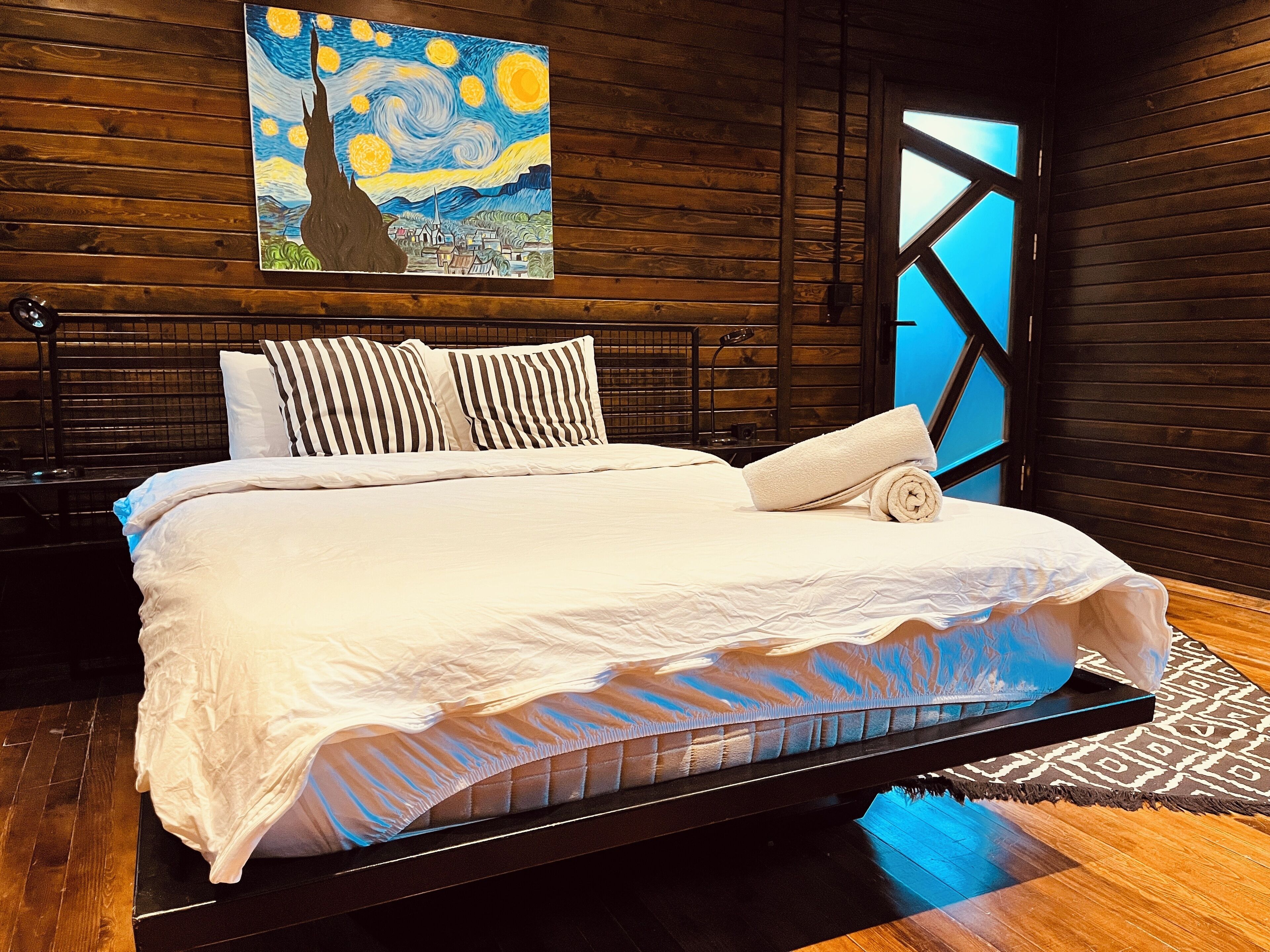 Un dormitorio moderno con cabecera de madera, un cuadro grande sobre la cama y vistas a través de una ventana de diseño geométrico.